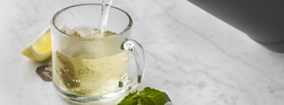 Water being poured into a mug to make a cup of tea. Next to it is a wedge of lemon and some mint leaves.